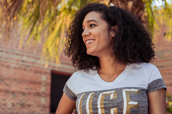 female student with curly hair and white/grey ucf shirt smiling into the distance (her right), while standing outside of a campus building under the trees
