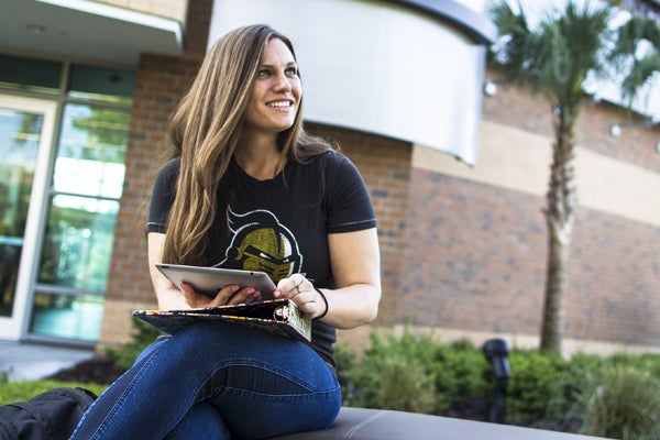 demale ucf student wearing black ucf shirt and blue jeans sitting outside holding book smiling off camera to the right