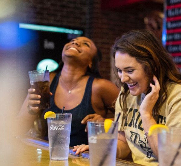 two female sutdents sitting at bar drinking water and soda, laughing and having fun on campus