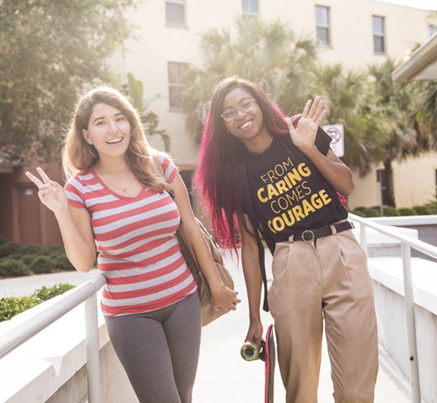 two female students standing and posing for the camera, leaning to their right waving and showing a peace sign with their fingers