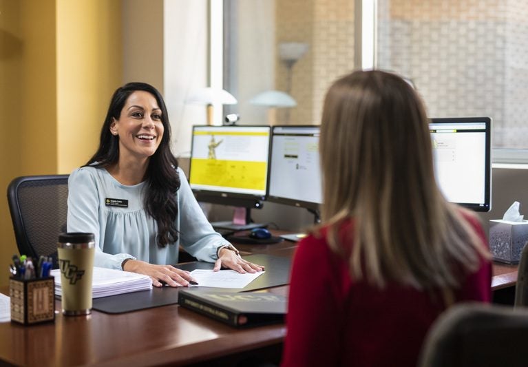 ucf employee sitting and desk, smiling and talking to female with back to camera