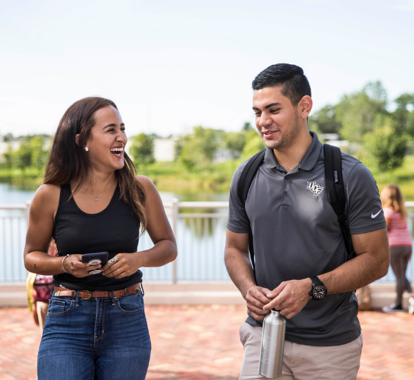 2 students walking on a brick courtyard near a pond. They are talking and laughing.