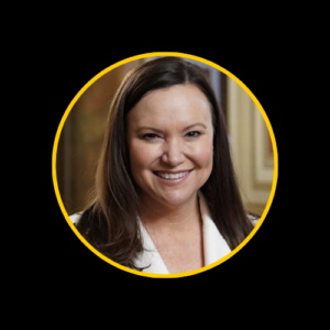 U.S. Senator Ashley Moody, a Caucasian woman with long brown hair, wearing a white blazer, smiling while standing in front of an American flag.