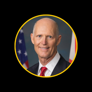 U.S. Senator Rick Scott, a Caucasian man with a bald head, wearing a dark suit, white shirt, and red tie, standing in front of American flags