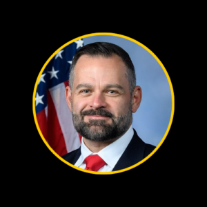 U.S. Representative Cory Mills, a Caucasian man with a full beard and short-cropped hair, wearing a dark suit, white shirt, and red tie, standing in front of an American flag