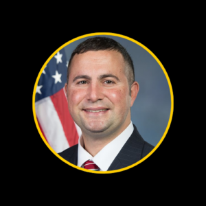 U.S. Representative Darren Soto, a Hispanic man with short dark hair, wearing a dark suit, white shirt, and striped tie, smiling in front of an American flag. 