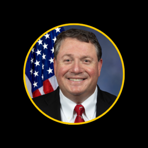 U.S. Representative Randy Fine, a Caucasian man with short dark hair, wearing a dark suit, white shirt, and red tie, smiling in front of an American flag.