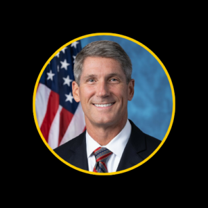 U.S. Representative Scott Franklin, a Caucasian man with short gray hair, wearing a dark suit, white shirt, and red tie, smiling in front of an American flag.
