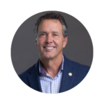Fred Kittinger, Senior Associate Vice President for Government and Community Relations at UCF, smiling in a professional headshot. He is wearing a blue blazer with a UCF pin and a light-colored checkered shirt, posed against a gray background.