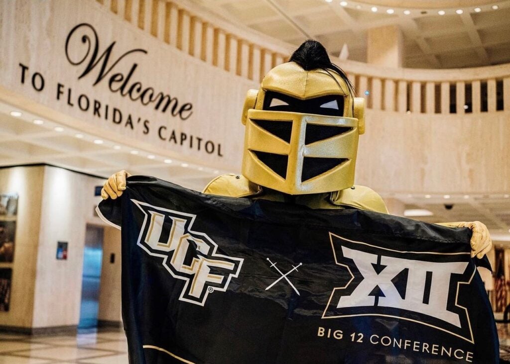 UCF’s mascot Knightro stands inside Florida’s Capitol holding a black flag featuring the UCF logo, crossed lances, and the Big 12 Conference emblem. The background displays a large sign reading 'Welcome to Florida’s Capitol