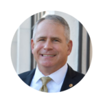 Eric "Tubby" Shwedo, Associate Vice President for Federal Relations at UCF, smiling in a professional headshot. He is wearing a dark suit jacket, white shirt, and gold tie, with a UCF pin on his lapel, standing outdoors in front of a light-colored building