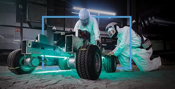 wo people in protective suits work on a rover in the Exolith Lab