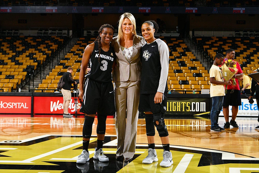 A white woman in a beige suit stands between two black players on a basketball court with empty seats in the background.