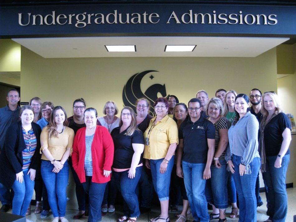 group of ucf's undergraduate admissions staff standing in front of pegasus logo and below a black sign with gold lettering 'undergraduate admissions'