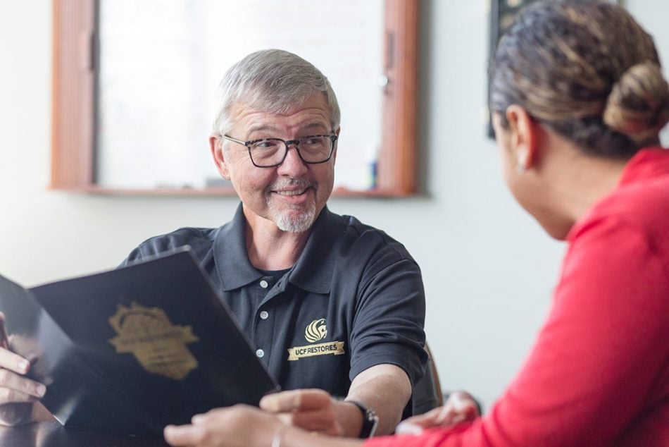 grey haired man with glasses and pegasus logo collared shirt talking to ethnic woman to his left