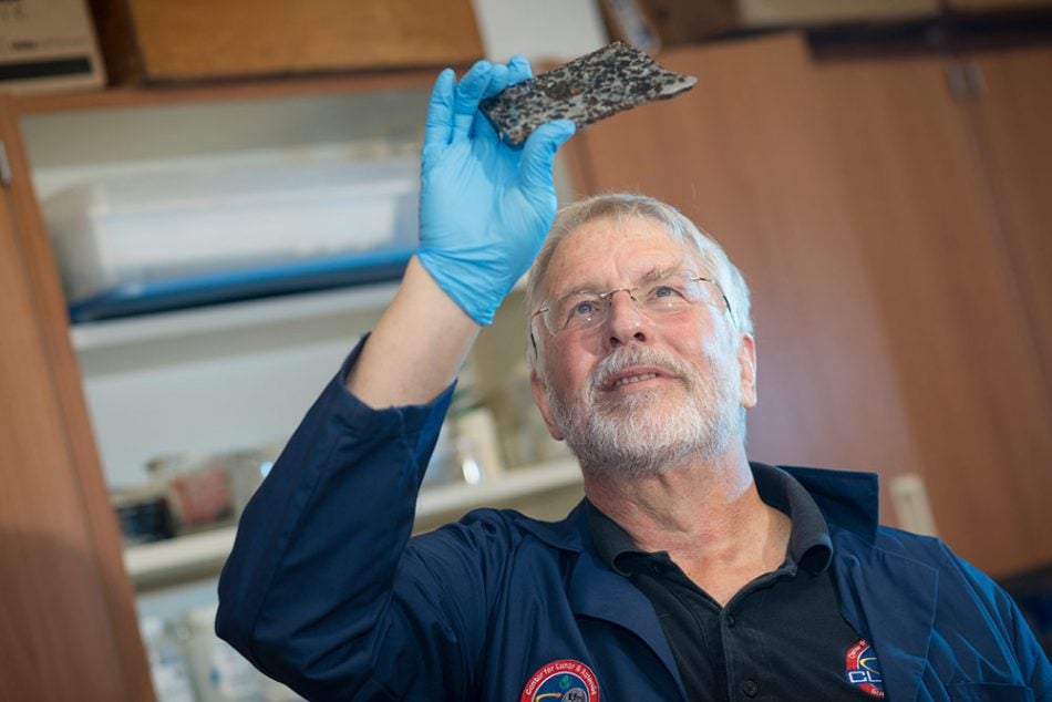 grey haired professor with glasses holding up a piece of asteroid with blue latex glove on his right hand