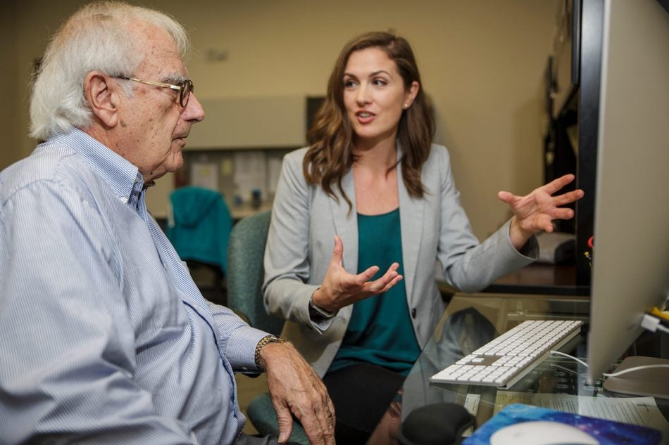 brunette woman in green shirt and grey blazer talking excitedly to an elderly man with white hair, glasses, and blue button down siting in front of a computer.