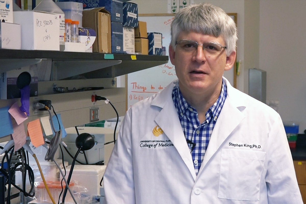 A white man with silver hair in white coat stands in lab with lots of equipment in the background.