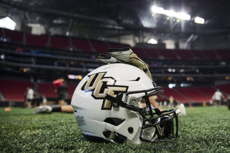 white football helmet with black and gold UCF written on the side. gold gloves placed on top of the helmet. the helmet is sitting on the grass of a football stadium while players practice in the background