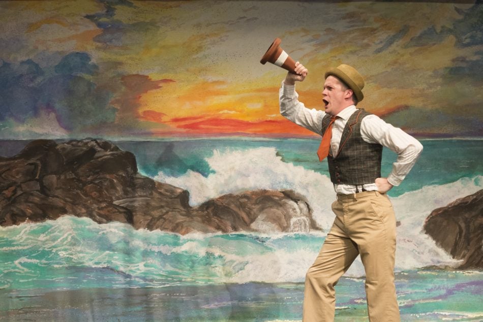 man in khaki pants, white long sleeve collared shirt, dark green vest, orange tie, wearing a tan hat is pretending to yell into a small traffic cone. standing in front of a mural of water hitting rocks in front of a sunset.