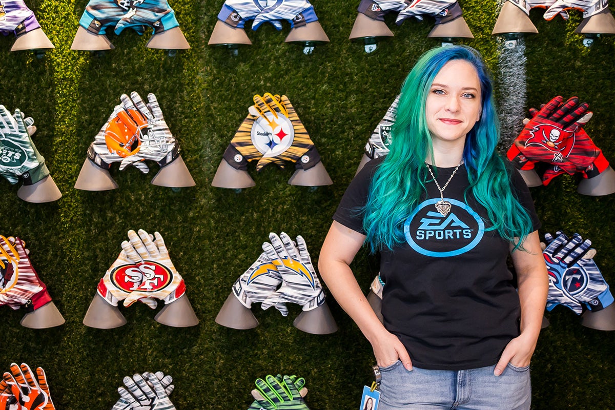 Young woman with blue/green long hair wearing a black t shirt and blue jeans poses with her hands in her front pockets in front of a grass-green wall featuring hand gloves of various NFL teams