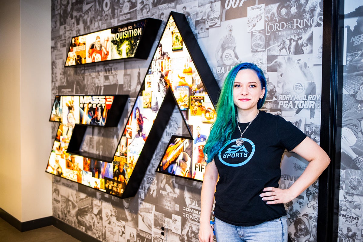 Young woman with blue/green long hair wearing a black t shirt and blue jeans poses with her left hand on her hip in front of black and white wall with multi-color EA sports logo