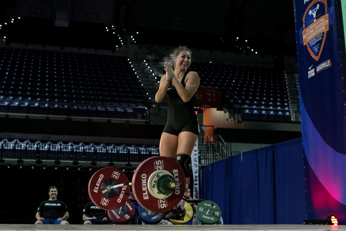 Young adult woman claps and smiles on stage with a bench press bar and weights at her feet