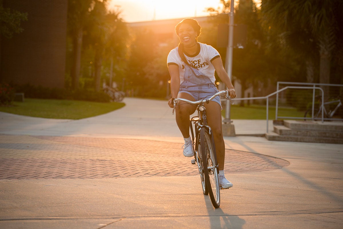 Young woman wearing a white t shirt and jean, short overalls, pedals a silver bike with soft light from sun setting behind her