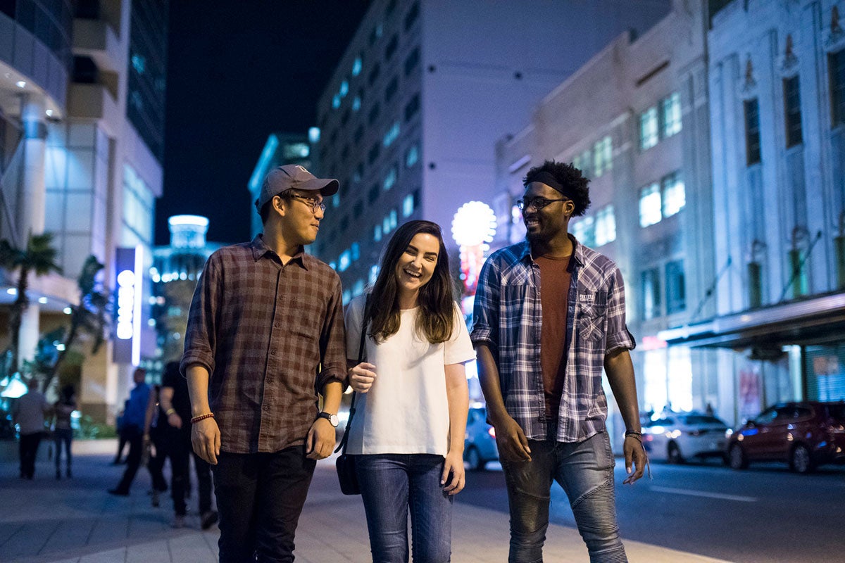 A young adult female with brown hair, white t shirt and blue jeans, walks downtown at night flanked by two men, one wearing a baseball cap, flannel shirt and jeans and the other wearing glasses, a black head band, blue open button flannel with a red-brown shirt underneath and blue jeans.