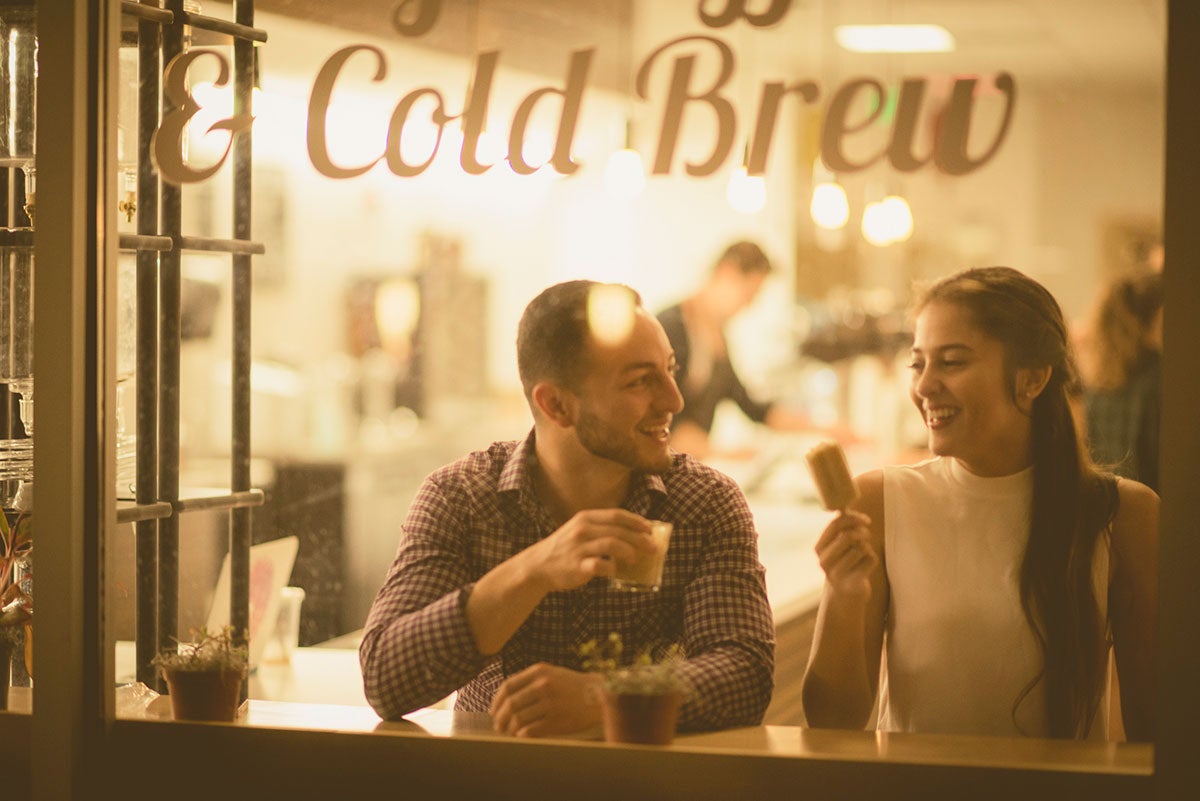 Young man and woman sit inside Pop Parlour storefront at a window. Man with a dark hair and long sleeve shirt holds a small coffee cup and smiles at woman in a white sleeveless shirt holding a popsicle.