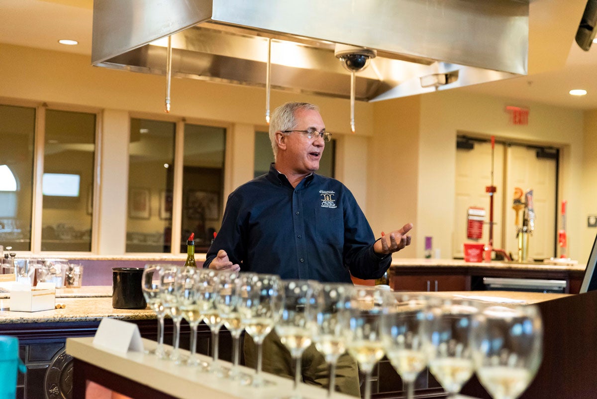 An older man with gray hair wearing a navy blue shirt gestures in front of a row of wine glasses