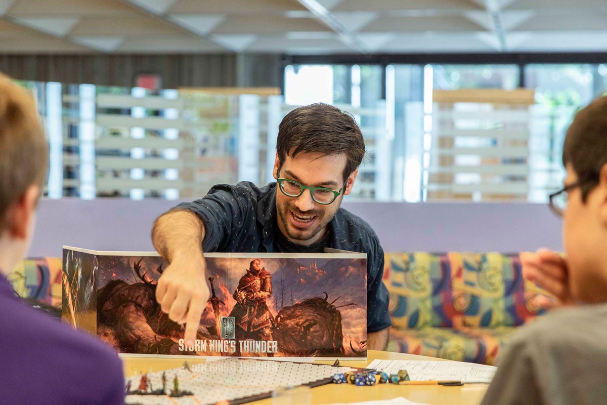 A man with dark hair and wearing glasses reaches over a book standing upright in front of him and points to a piece on a gaming board.