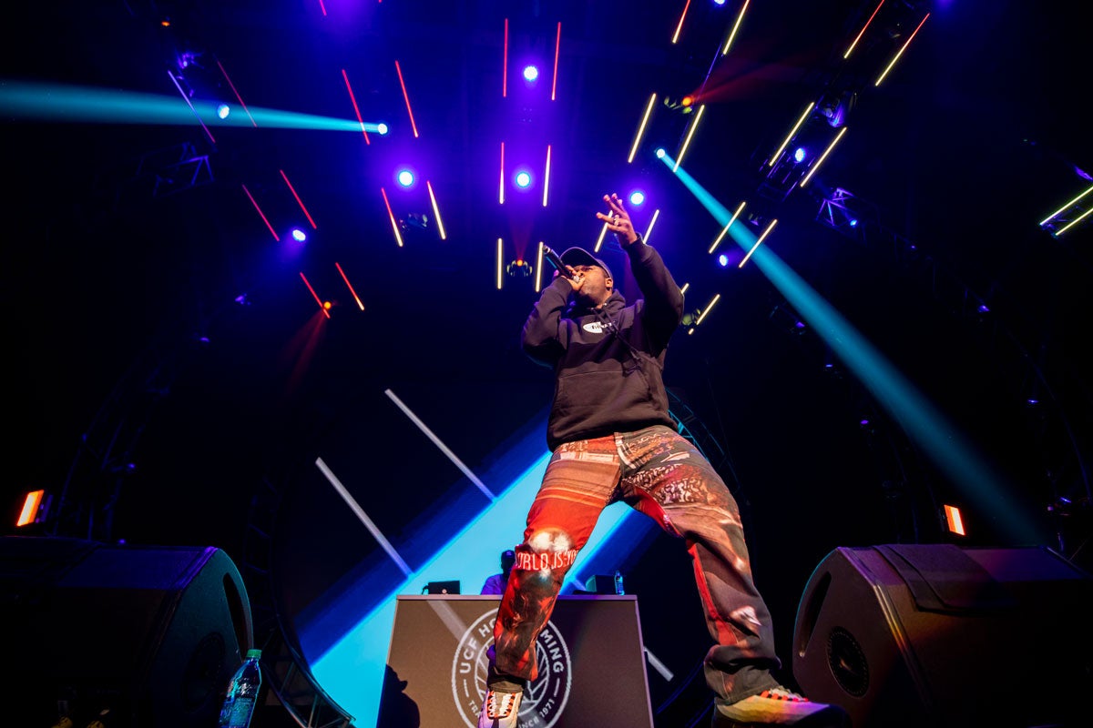 a man wearing multicolor red pants and a black long sleeve shirt holds a microphone with blue lasers shining around him on stage