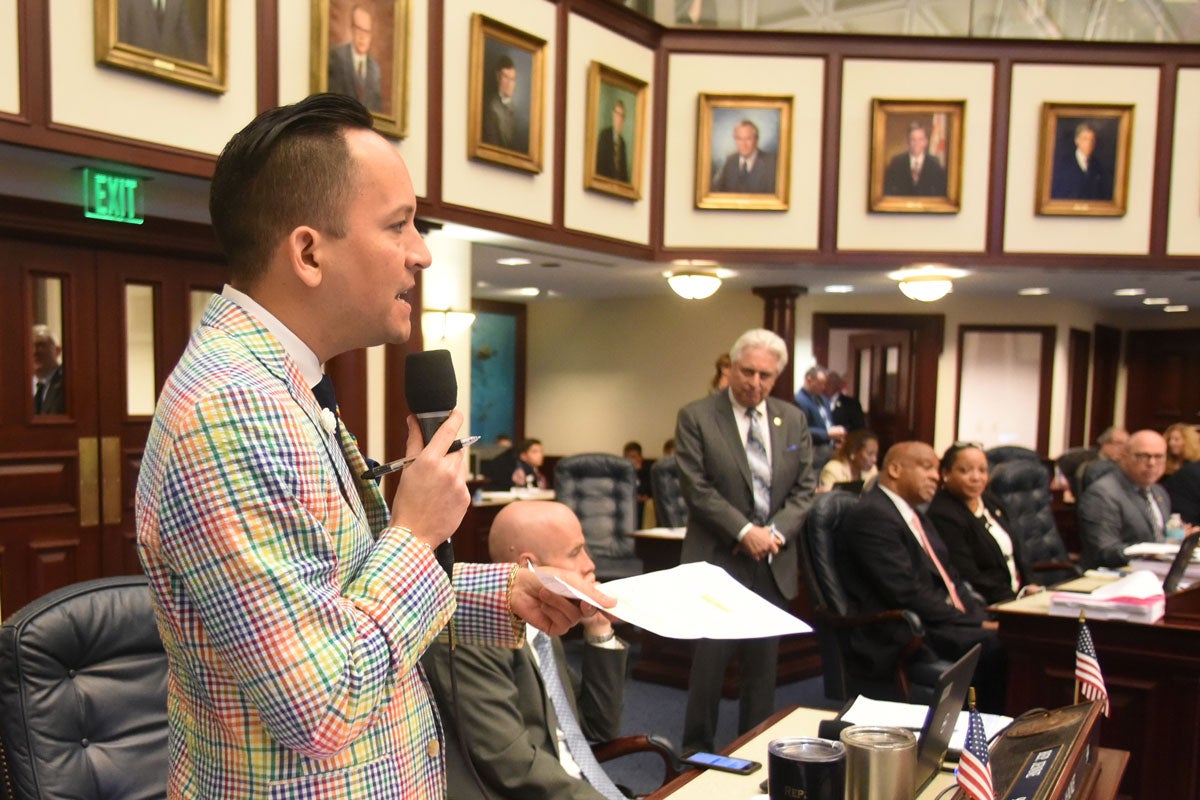 A man wearing a multi-color suit stands in a legislative hall while holding a microphone with his right hand to address his colleagues.