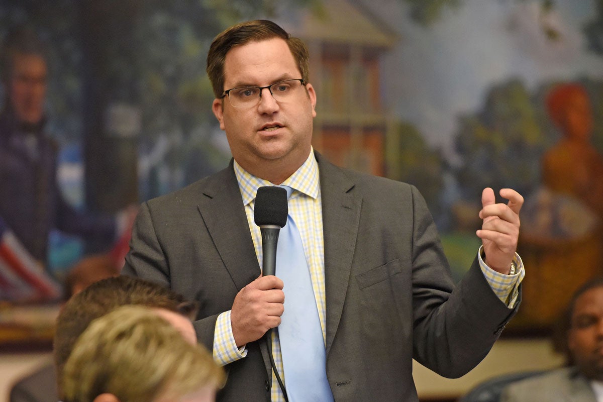 A man with brown hair, wearing glasses and in a gray suite with a light blue tie and cream shirt holds a microphone