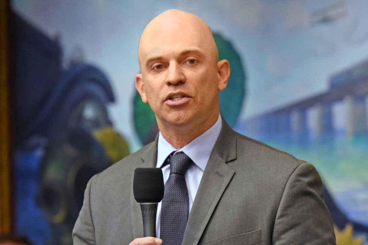 A bald man wearing a gray suite with a light blue shirt and dark blue tie holds a microphone