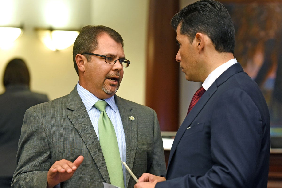 A man with short brown hair wearing glasses in a suit with a green tie talks to another man wearing a blue suit 