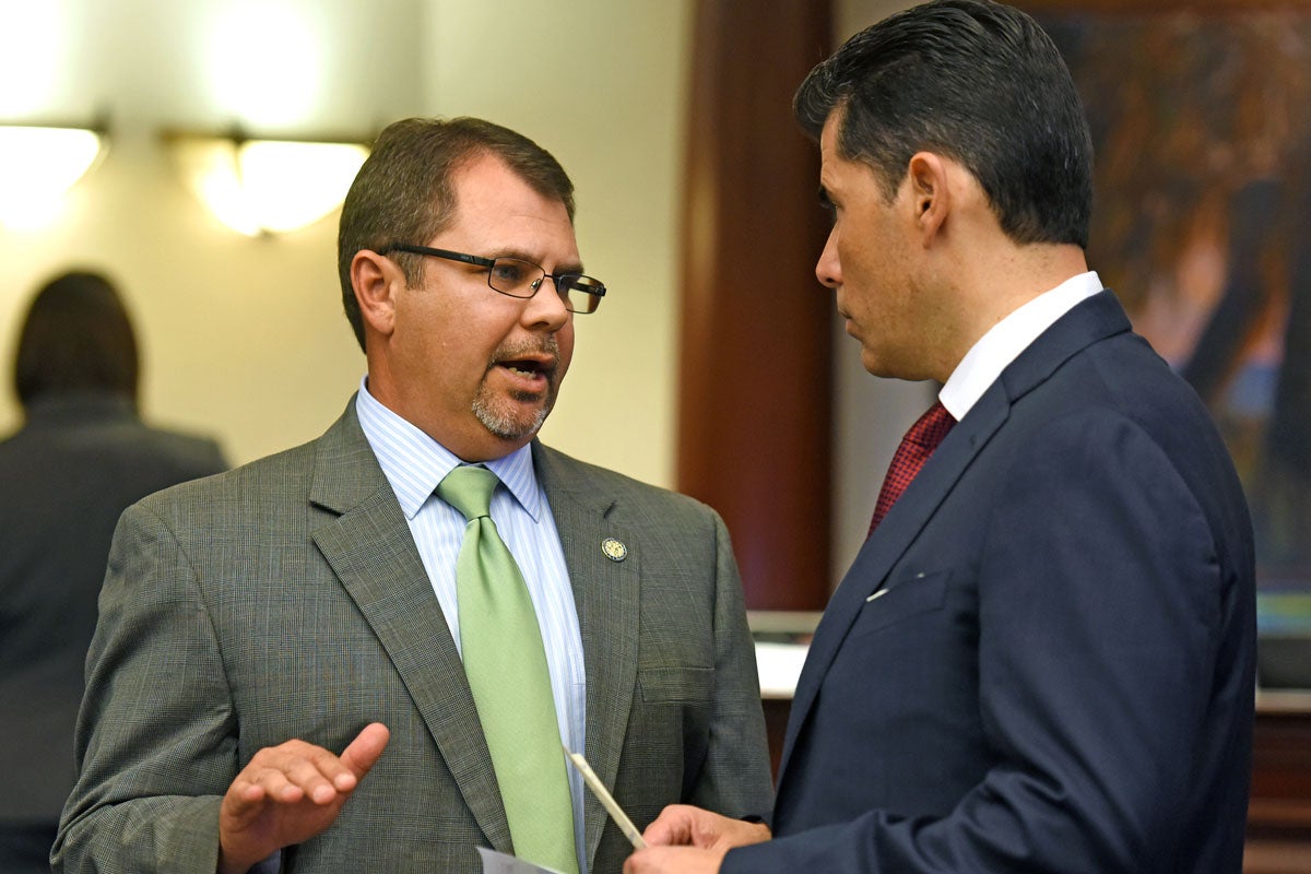 A man with short brown hair wearing glasses in a suit with a green tie talks to another man wearing a blue suit