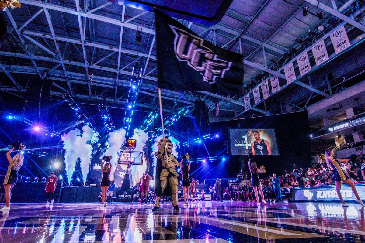 UCF mascot Knightro waves black UCF flag on court at CFE Arena with cheer team around him