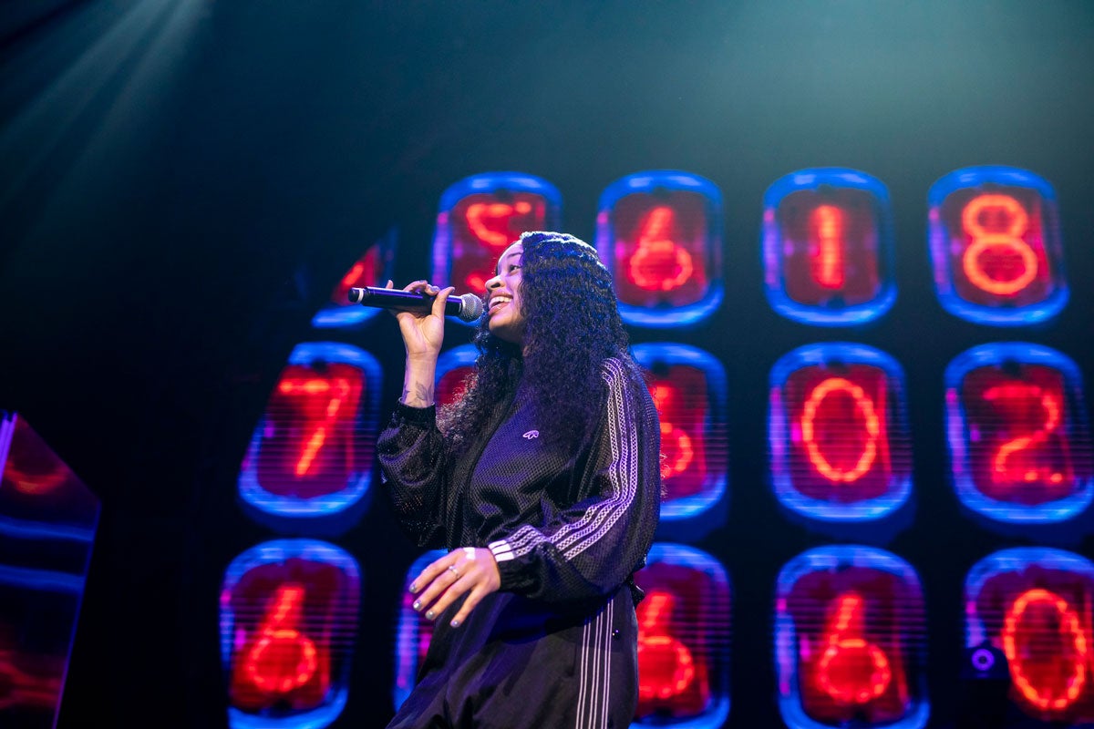 a woman in a black track suit sings in front of neon red numbers 