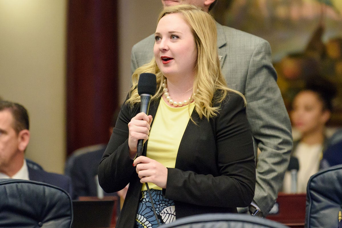 A young woman with blonde hair wearing a yellow shirt and black blazer stands holding a microphone