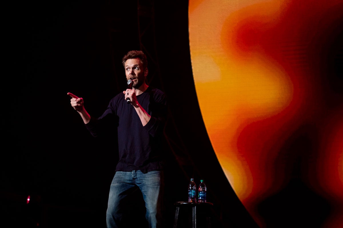 Comedian Joel McHale, wearing a black long sleeve shirt and jeans, holds a microphone and points to the crowd at CFE Arena