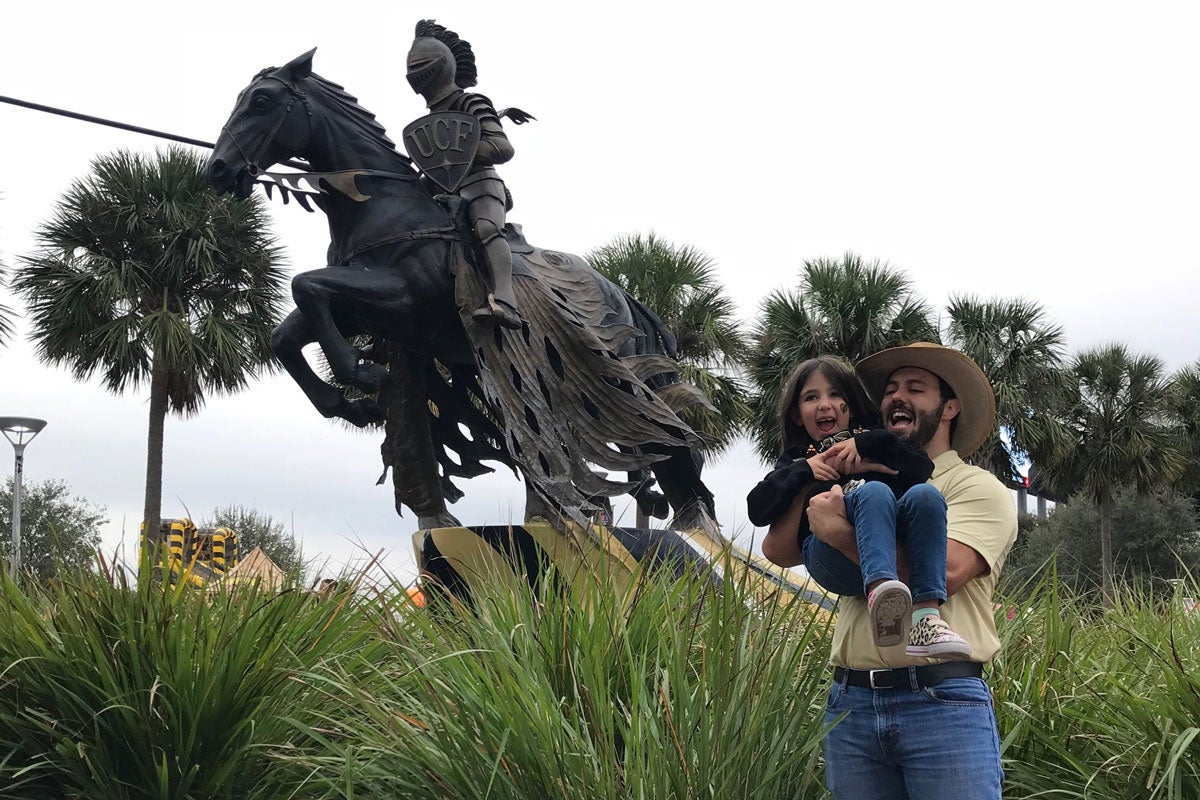A man wearing a straw hat, yellow t shirt and blue jeans cradles a small child in front of a bronze statue of a knight holding a lance while riding a horse