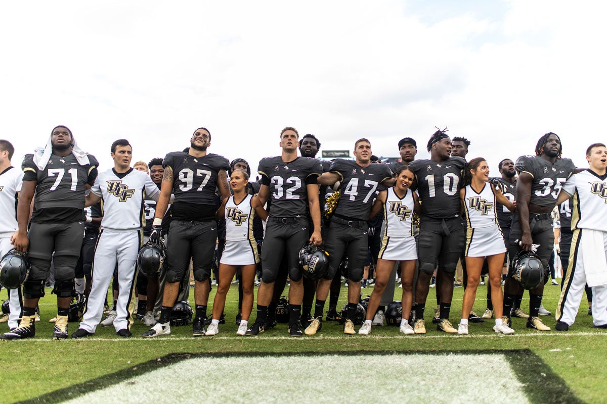 Football players and cheerleaders are linked arm in arm in the end zone of a football field