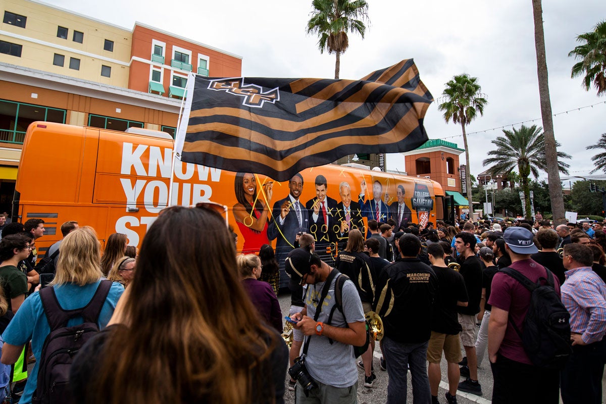 Black and Gold UCF flag waves in front of orange bus with a crowd around it