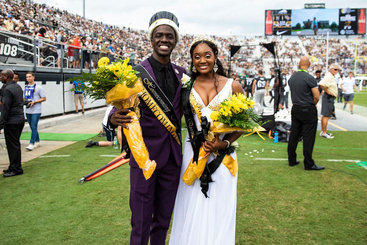 a man in a black suit wearing a king crown and a woman in a white gown both hold flowers and stand on a football field as Homecoming king and queen