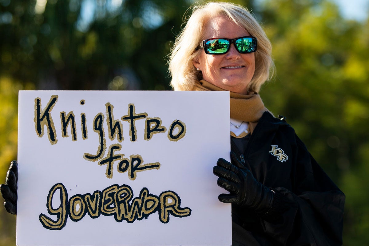 Blonde woman wearing sunglasses holds sign that says Knightro for Governor