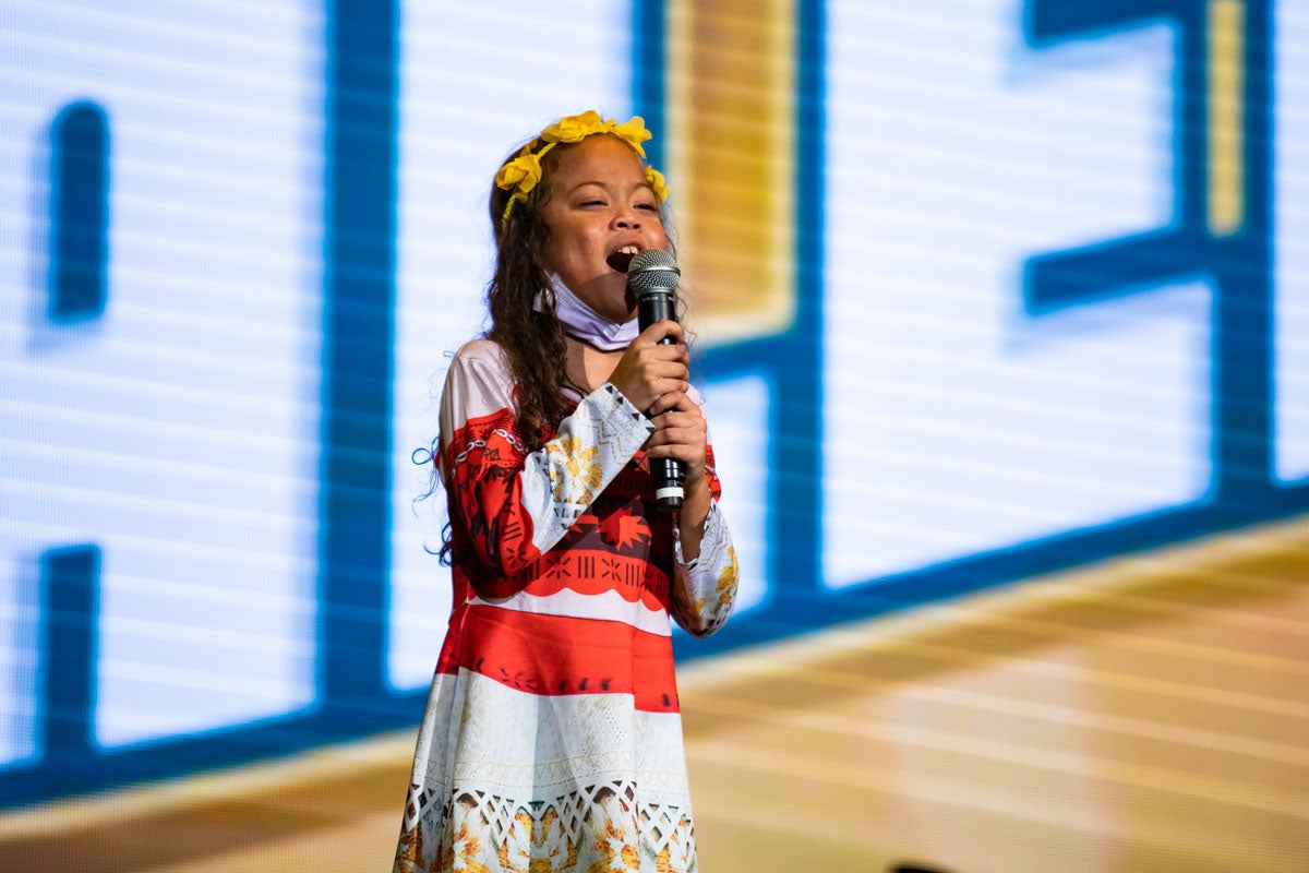 A young girl with a yellow headband and red and gray dress sings into a microphone