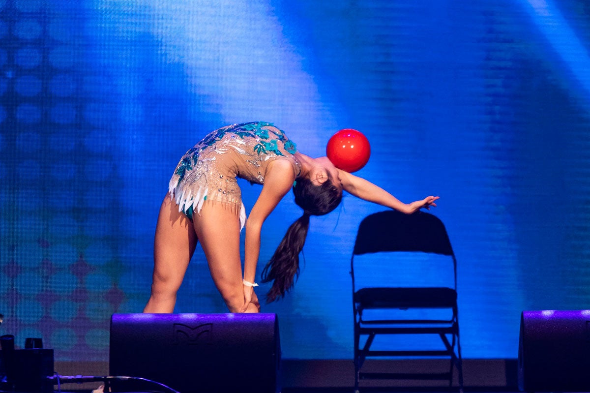 A woman with a black pony tail does a standing back bend while balancing a red ball on her outstretched arm
