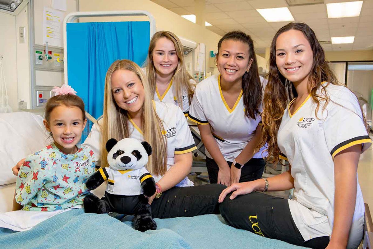 four UCF nursing students give a stuffed panda bear to a girl wearing a green gown in a hospital bed.
